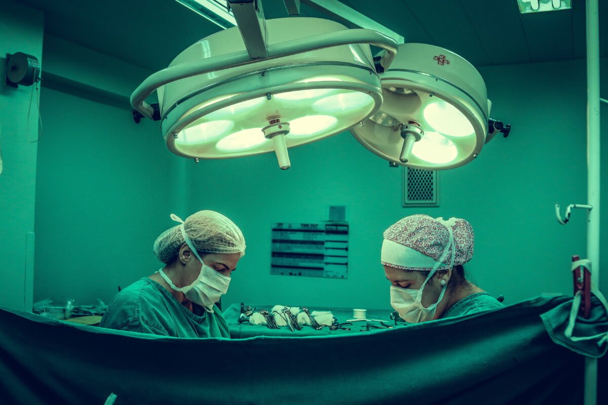 Two surgeons stand in an operating room and operate. You can only see her body up to the shoulder, a cloth hangs in front of it.