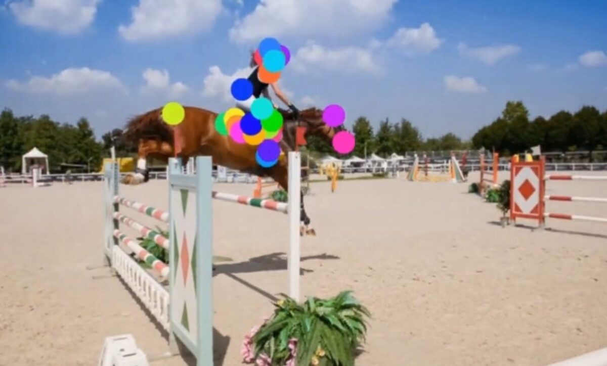 A show jumper jumps over an obstacle with her horse. The woman and the horse have colored dots on their heads and bodies, which the AI uses to track their movements.