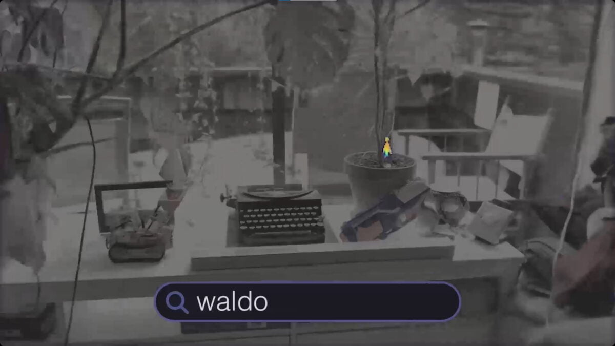 A 3D NeRF scene of a desk. A small "Where is Waldo" figure stands in a flower pot. The voice command in a search mask "Waldo" can identify this figure and then zooms in.