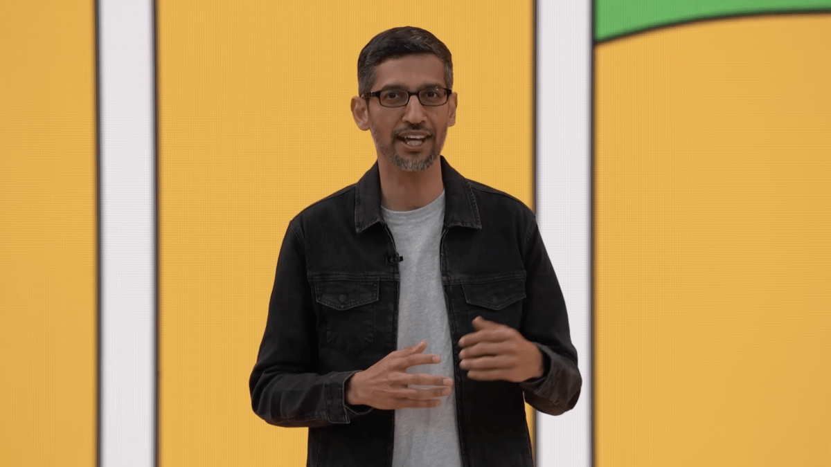 Google CEO in front of a yellow video screen at a lecture, speaking into the camera.