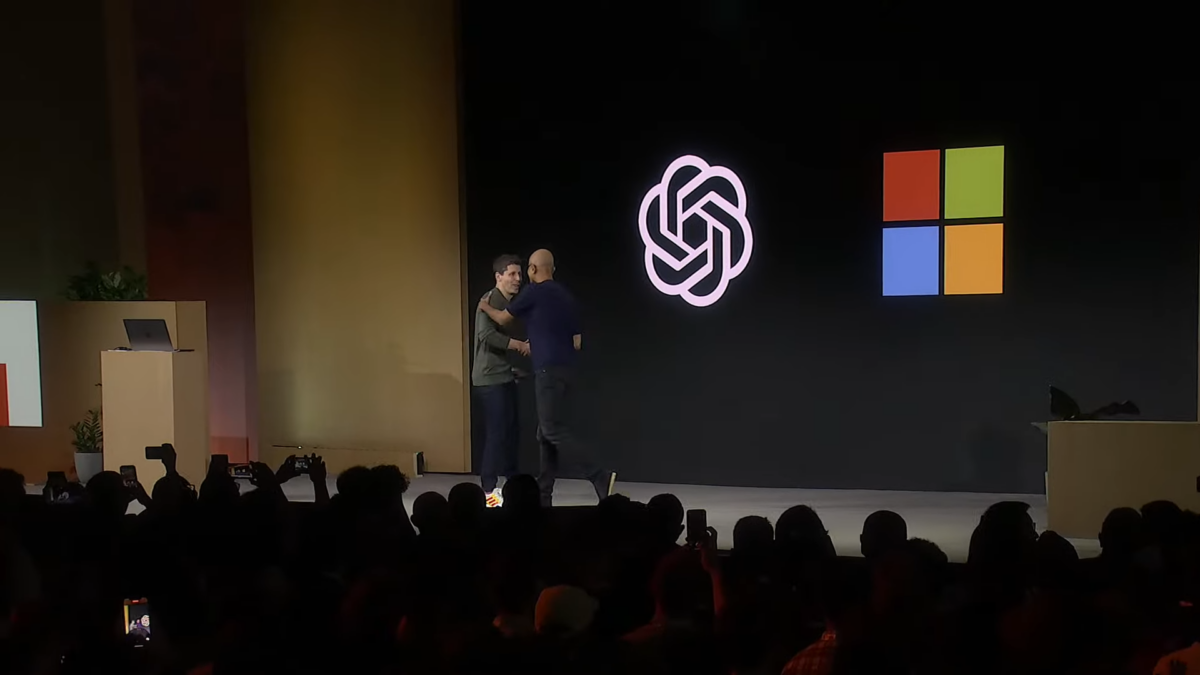 Sam Altman from OpenAI and Microsoft CEO Satya Nadella shake hands on stage at the OpenAI Dev Days. On a screen in the background are the OpenAI and Microsoft logos.