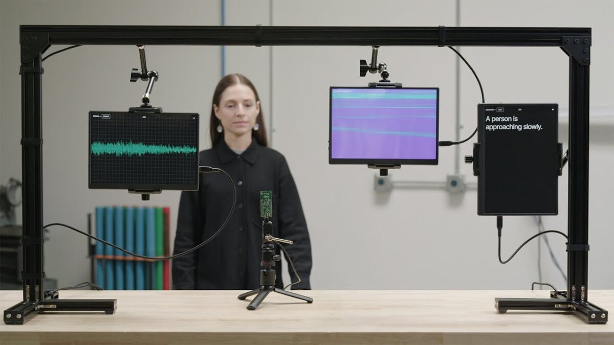 A woman stands in the background between two mounted screens. The left screen displays a waveform, possibly audio levels, on a grid background. In the center, there's a circuit board mounted on a tripod with cables connected to it. The right screen shows text reading "A person is approaching slowly." The setup suggests a demonstration of some sensor technology detecting and responding to the presence of a person.