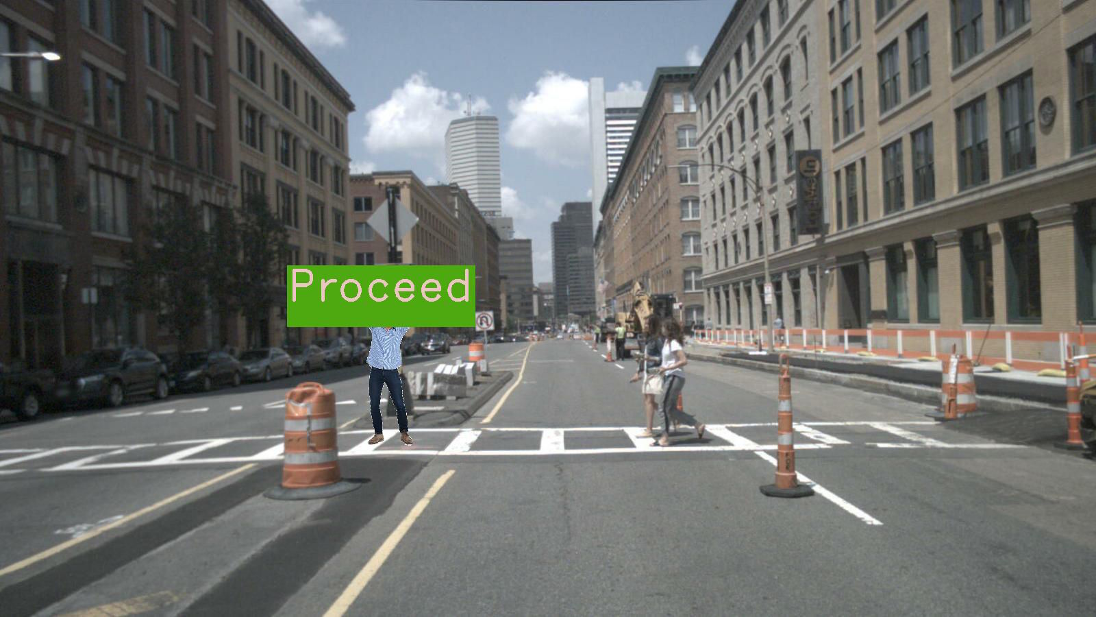 Man in construction site holding green sign saying "Proceed" while pedestrians cross a city street.
